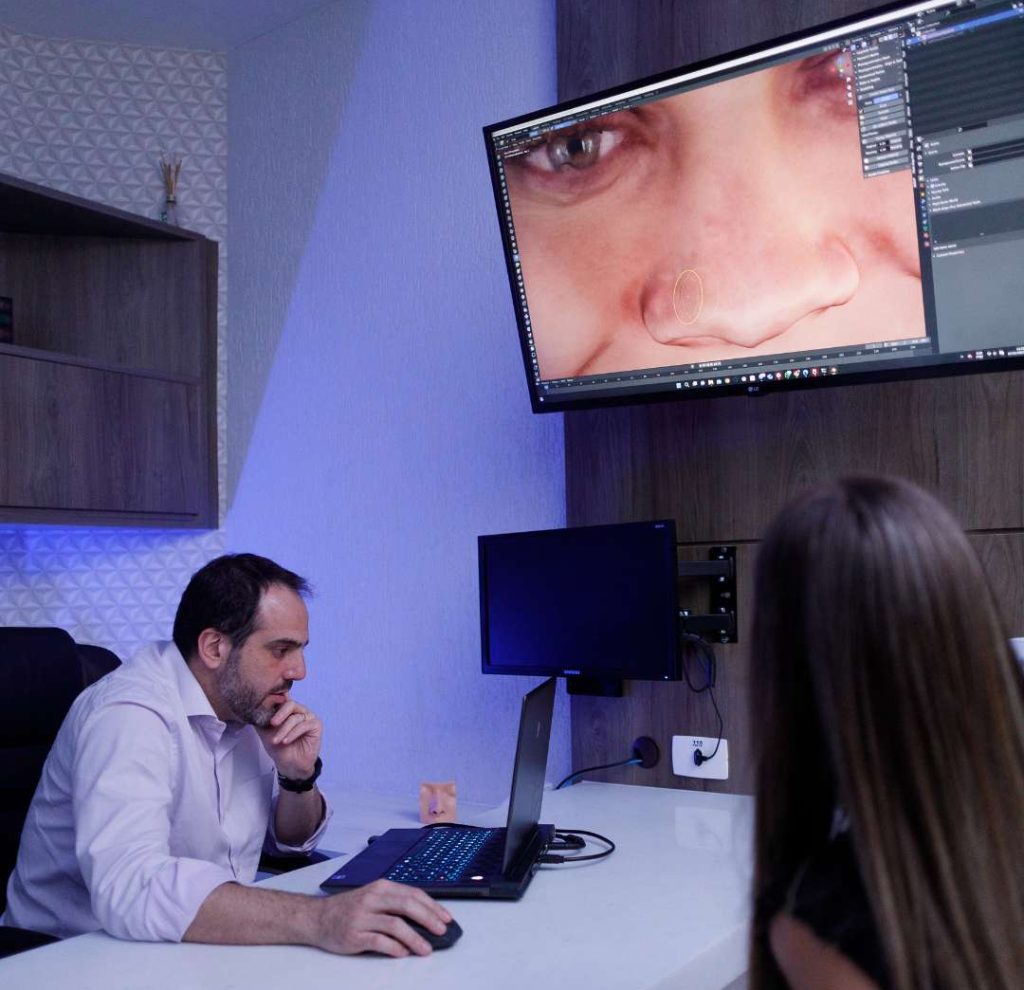 Médico concentrado realizando trabalho de simulação em seu computador sobre a mesa enquanto do outro lado da mesa mulher presta atenção na imagem de seu rosto projetada na tela.
